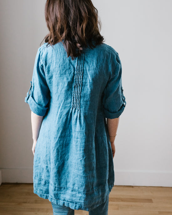 A woman seen from behind, wearing a Regina in Bleach Indigo Twill tunic dress from CP Shades and standing in front of a plain wall.