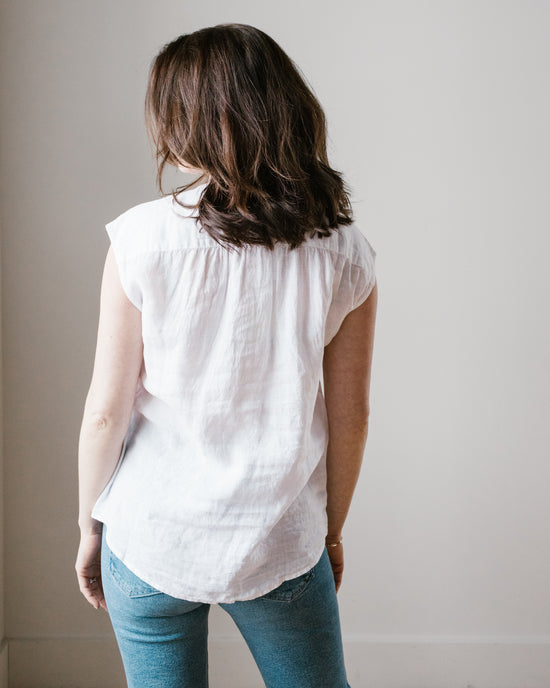 A woman with medium-length hair, wearing a Hartford Cleo Shirt in White and blue jeans, stands facing a wall.