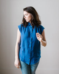 A woman in a relaxed silhouette, wearing a blue sleeveless button down Hartford Cleo Shirt in Mykonos and gray pants, standing against a plain background, looking to the side.