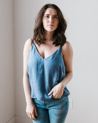 A woman wearing a Bella Dahl Adj Strap V Neck Cami in Mykonos Blue and jeans standing against a white wall.
