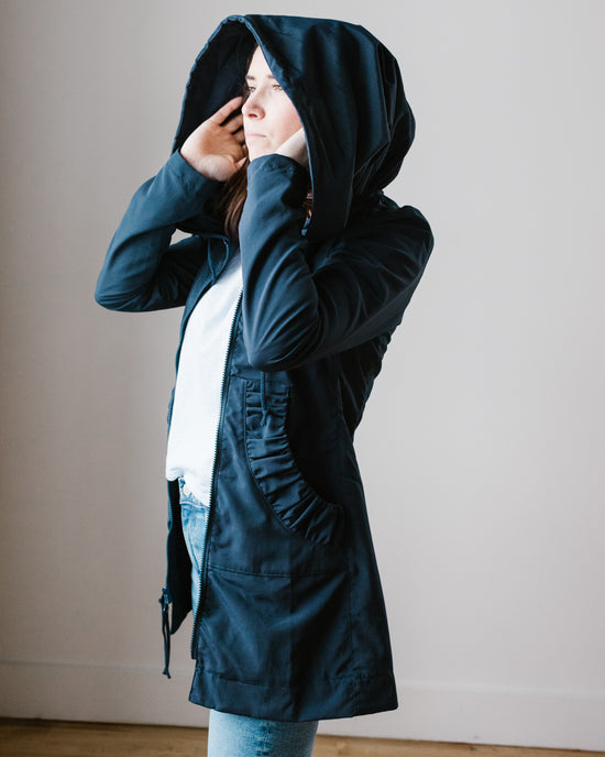 A person in a Prairie Underground Mid Raincoat in Navy, hooded coat covering their face with one hand.