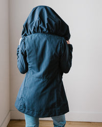 A person standing with their back to the camera, wearing a Prairie Underground Mid Raincoat in Navy and covering their head with the hood.
