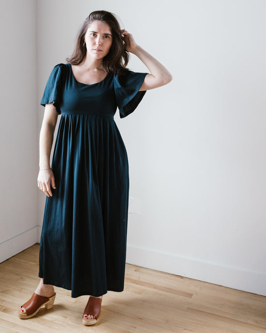 A woman in a blue Demylee Elana Dress in Navy made of pima cotton jersey, standing against a white wall, touching her hair.