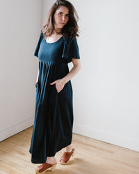 A woman in a pima cotton jersey Demylee Elana Dress in Navy and open-toe heels stands in a room with a neutral expression.