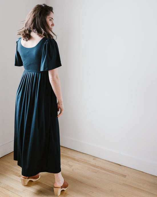 Woman in a dark blue pima cotton jersey maxi dress, known as the Demylee Elana Dress in Navy, standing beside a white wall, looking over her shoulder.