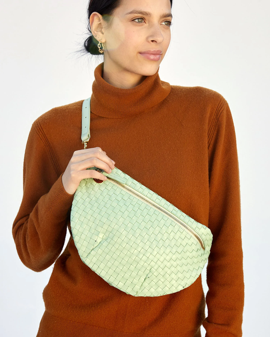 Woman in a brown turtleneck sweater holding a Clare V. Grande Fanny in Mist Woven Checker bag, against a plain background.