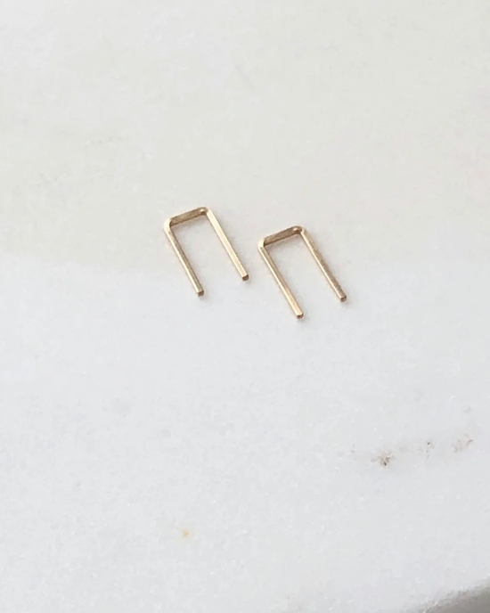 Two small metal staples on a white surface, replicating Token Jewelry's Mini Staple Earrings in 14K Gold Fill.