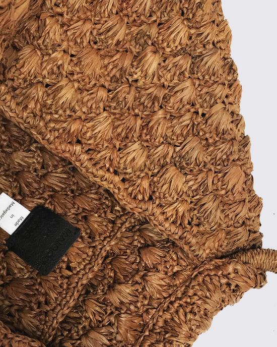 A close-up of a textured woven Clementine Bag in Sucre Brown with a clothing label from Maison N.H. Paris, crafted from natural rafia.
