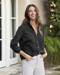 A smiling woman in a black Joedy in Black Superluxe Blouse and white pants standing by a door.