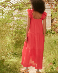 Woman in the Great's The Dove Dress in Tart, a lightweight cotton, coral pink dress with ruffled shoulders, standing in a greenhouse.