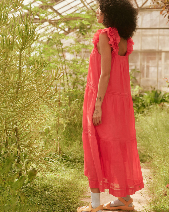 Woman in a pink dress with ruffled shoulders standing in a greenhouse wearing The Dove Dress in Tart by the Great.