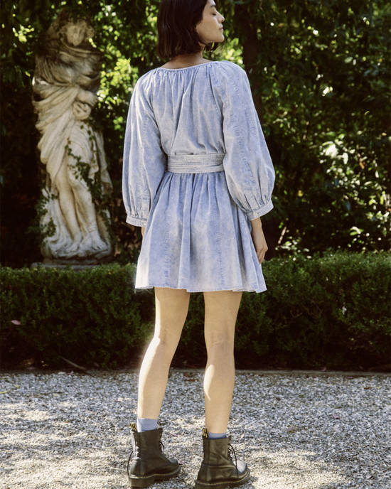A woman in The Great's Coast Walk Dress in Hand Dyed Mottled Wash, featuring voluminous sleeves, stands in a garden, with a sculpture visible in the background.