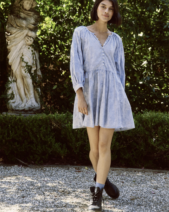 A young woman in a light blue The Great Coast Walk Dress in Hand Dyed Mottled Wash with voluminous sleeves and black boots standing in a garden with a statue in the background.