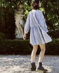 Person in the Great's The Coast Walk Dress in Hand Dyed Mottled Wash and black boots standing in a garden with a statue in the background.