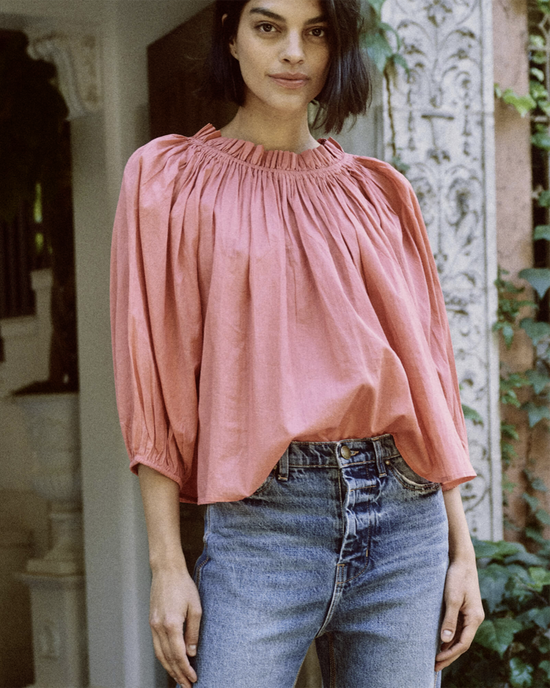 A woman wearing The Prim Top in Cherry Blossom by the Great with an elastic ruffled neckline and blue jeans standing in front of The Great Cherry Blossom backdrop.