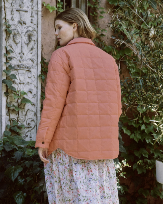 Woman in a The Reversible Cloud Puffer in Cherry Blossom w/ Rose jacket standing in front of a rustic wall by the Great.