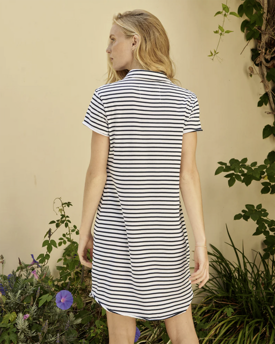 Woman wearing a striped V Neck Lauren Polo dress in White w/ British Royal Navy Stripe standing with her back to the camera in front of a plant-covered wall.