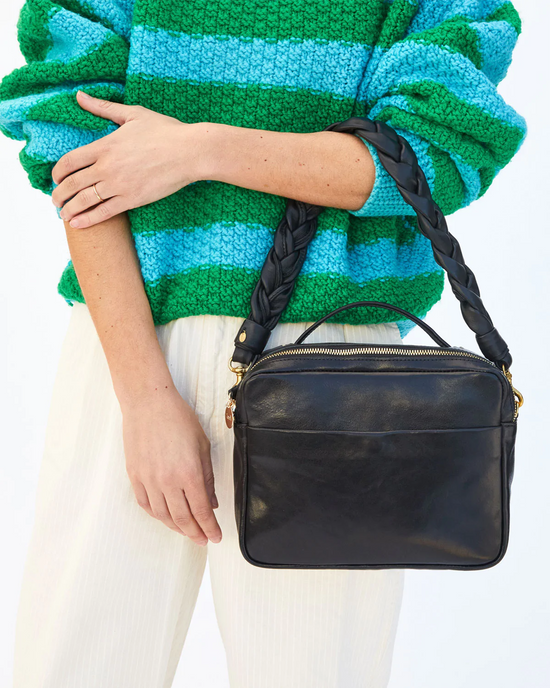 Woman holding a black Clare V. Braided Leather Shoulder Strap shoulder bag, complementing her colorful knitted sweater and white trousers.