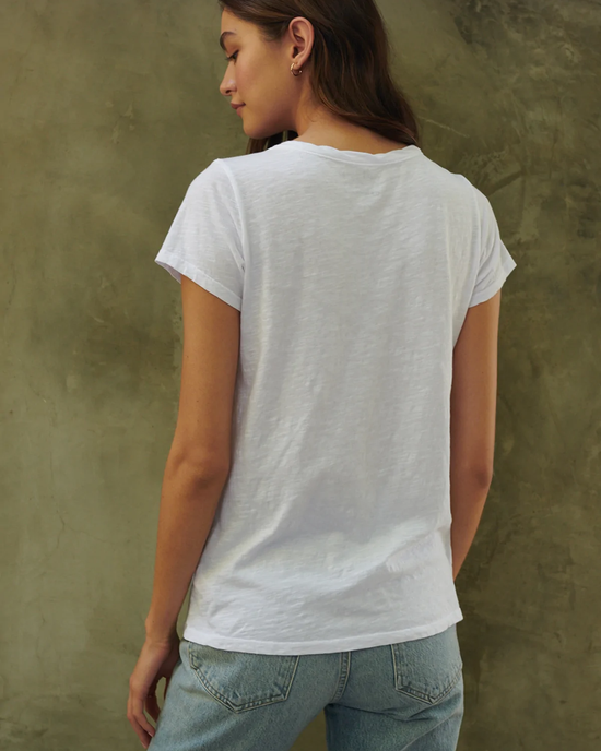Woman standing with her back to the camera, wearing a Velvet by Graham & Spencer Tilly Tee Shirt in White and blue jeans against a textured wall.