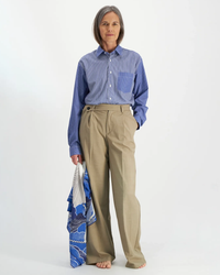 A woman standing against a white background wearing a blue-striped cotton/silk blend shirt, khaki trousers, and holding an oversized bandana patterned bag by Inoui Editions, barefoot.