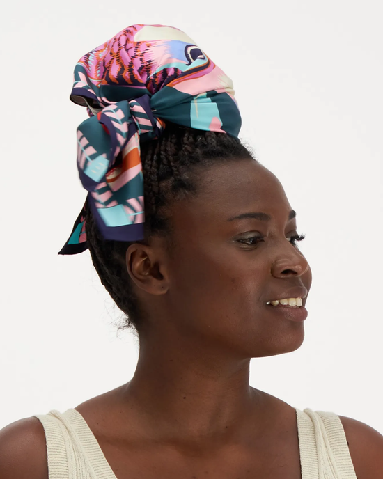 A woman wearing a colorful Inoui Editions Square 65 Toucan in Emerald silk scarf as a headwrap looks to the side with a thoughtful expression.