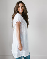 A person with long brown hair, wearing CP Shades' Cap Sleeve Regina in White Linen, paired with jeans, stands against a plain white background.