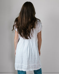 A woman with long brown hair stands facing a white wall, wearing a CP Shades Cap Sleeve Regina in White Linen over blue jeans.
