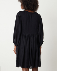 Woman standing with her back to the camera, wearing the Erin Shirred Waist L/S Dress in Black, a black cotton/silk dress with long sleeves and gathered waist detail by Velvet by Graham & Spencer.