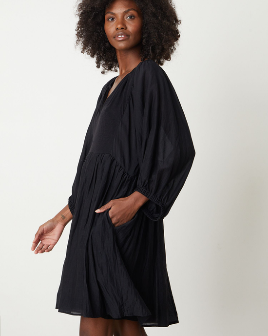 A woman in an Erin Shirred Waist L/S Dress in Black by Velvet by Graham & Spencer, with a relaxed fit and textured fabric, standing with one hand on her hip.