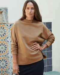 A woman with long brown hair stands by a mosaic-tiled wall wearing the Frank & Eileen Effie-L/S Funnel Neck Capelet in camel and a dark skirt, her left hand on her hip and a relaxed expression on her face.