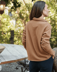 A woman with long brown hair, wearing the Frank & Eileen Patrick Henley in Camel Terry and dark pants, stands outdoors with her back to the camera beside a rustic wooden table, surrounded by greenery and soft sunlight.